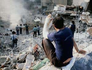 Person with arm in cast looking down on an expanse of rubble, smoke and twisted wires