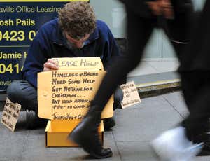 Man sitting on pavement holding a cardboard sign with downcast head