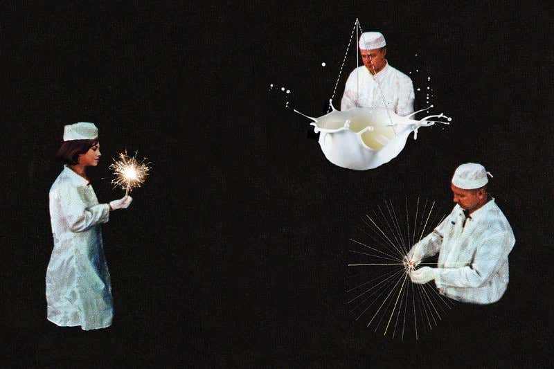 People in kitchen-workers' white outfits, one holding a sparkler, one looking at an enlarged splash in white liquid, one holding some kind of particle collision trail set