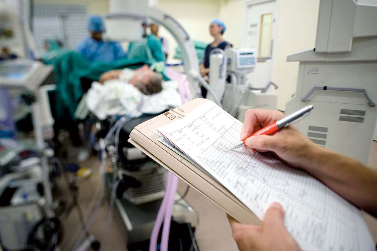 A doctor takes notes on a clipboard as a patient is receiving treatment in the background