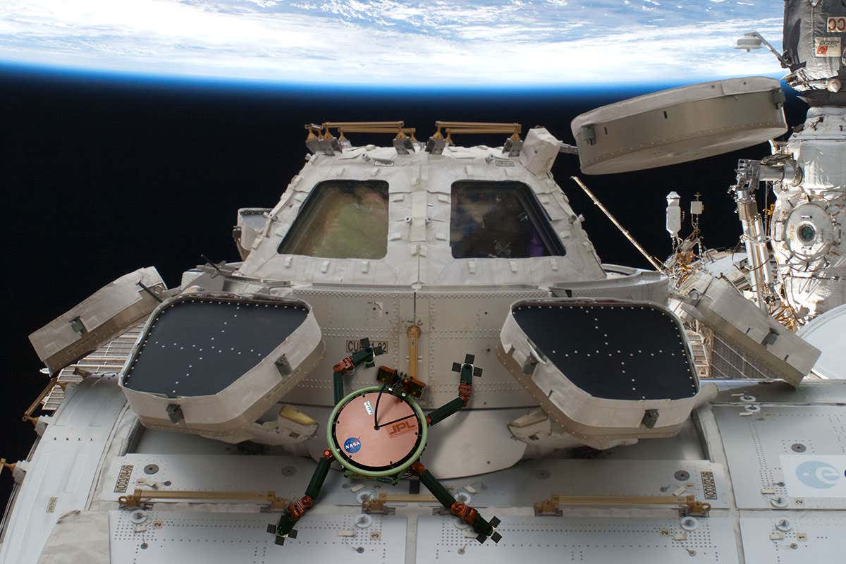 An artist's impression showing a small robot with four legs and gecko-like feet crawling over the outside of a spacecraft, with the surface of Earth visible over the top of the page