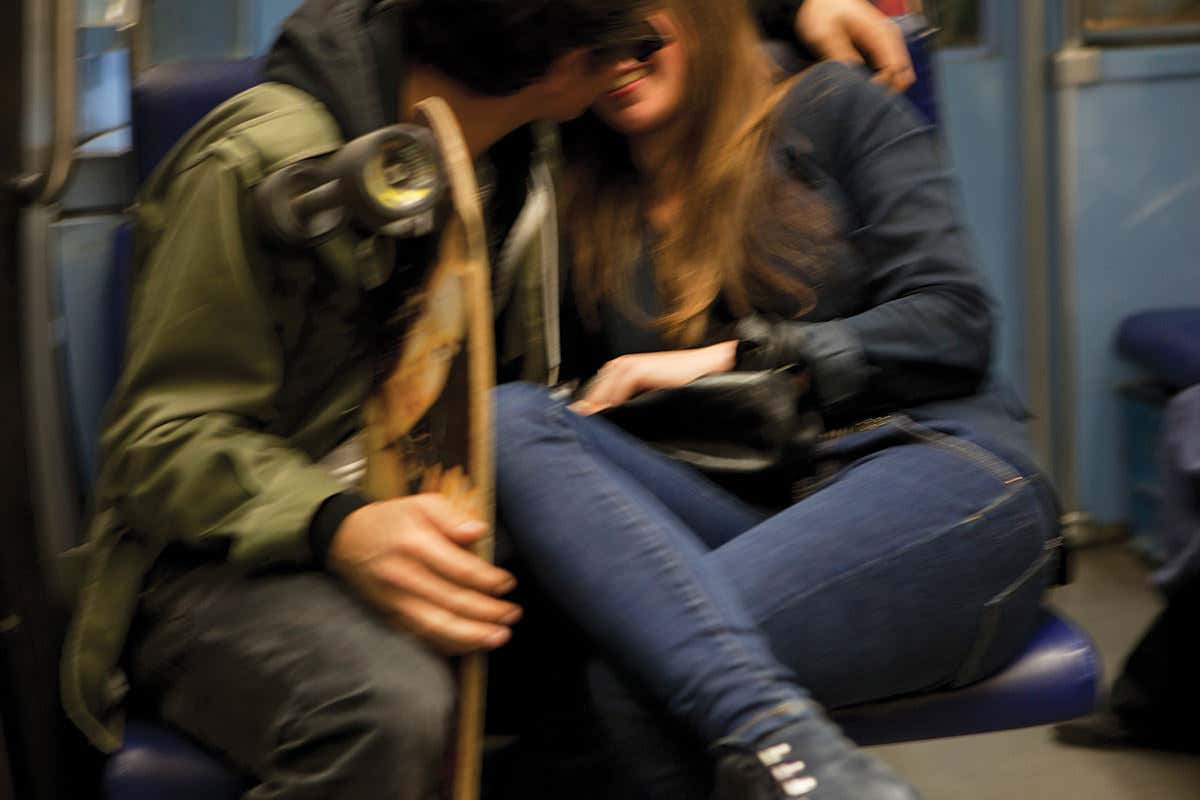 A young couple sitting next to each other, arms entwined. She's smiling as he leans in like they might kiss