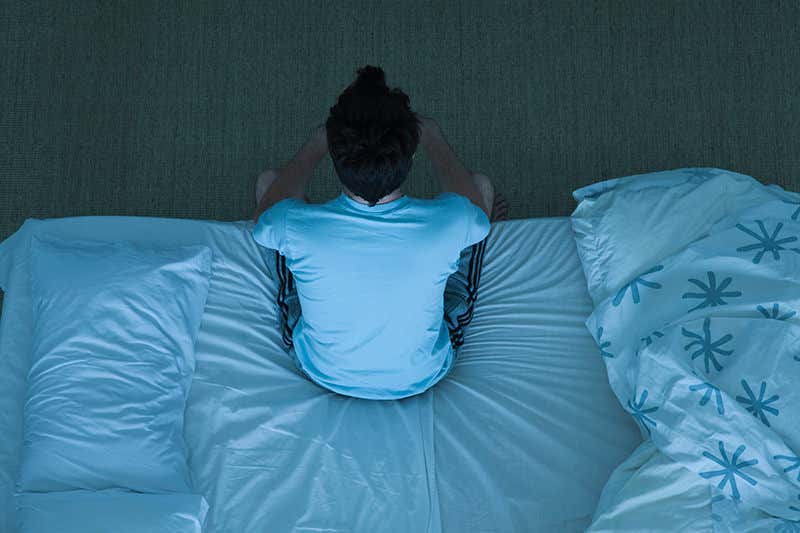 A person sits on the edge of a bed looking dejected
