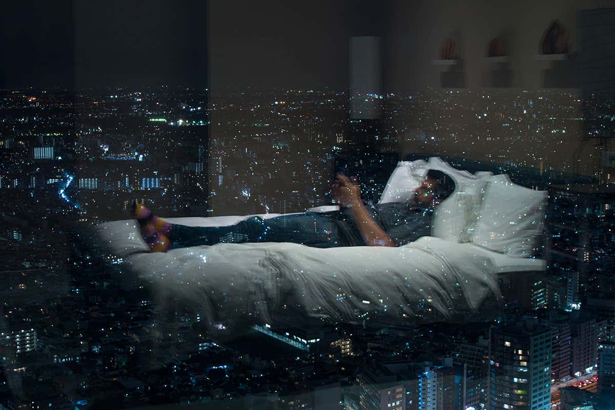 A man lying on a hotel bed with city lights all around through the windows