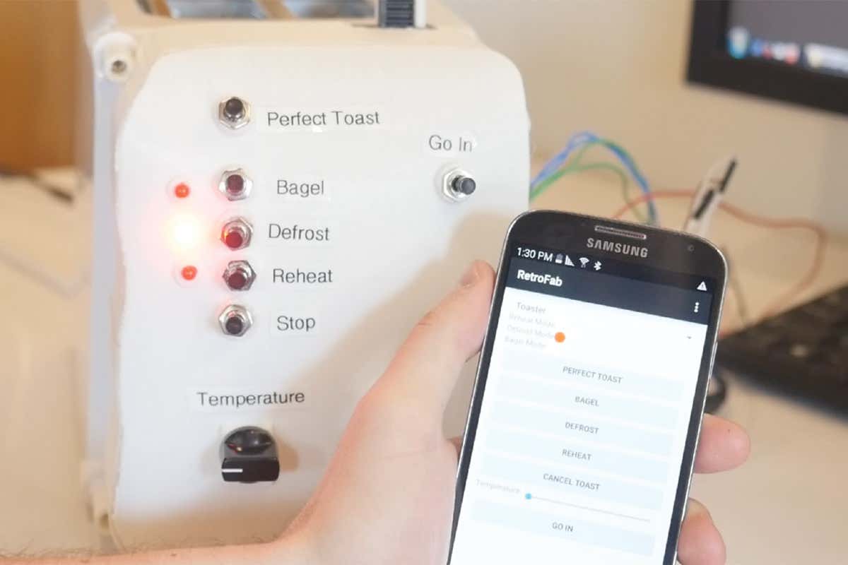 A new set of buttons attached to the end of a toaster, with a smartphone screen in someone's hand in the foreground demonstrating the controls for the toaster