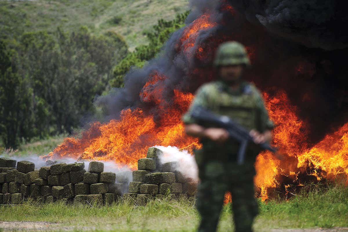 Armed soldier stands guard with rectangular packs ablaze in the background
