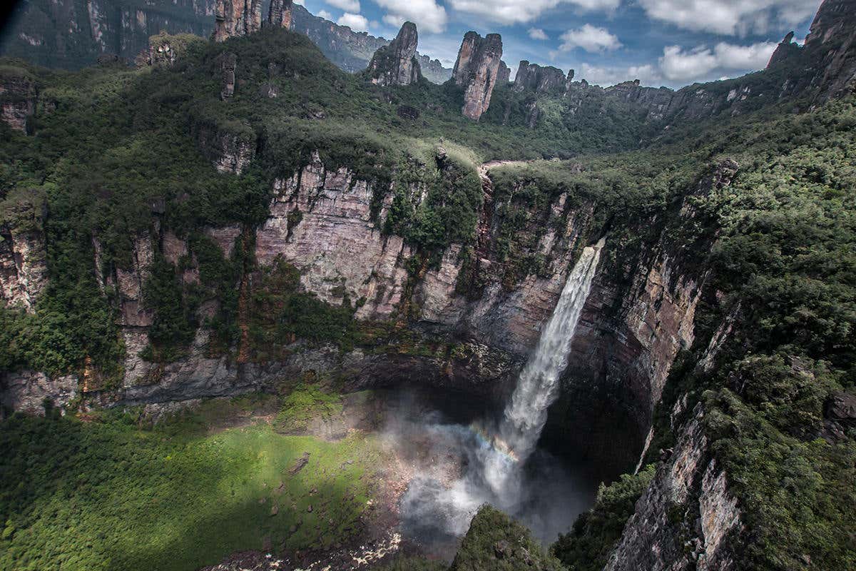 A spectacular landscape of cliffs with a waterfall plunging down to a crater and rocky pinnacles in the background