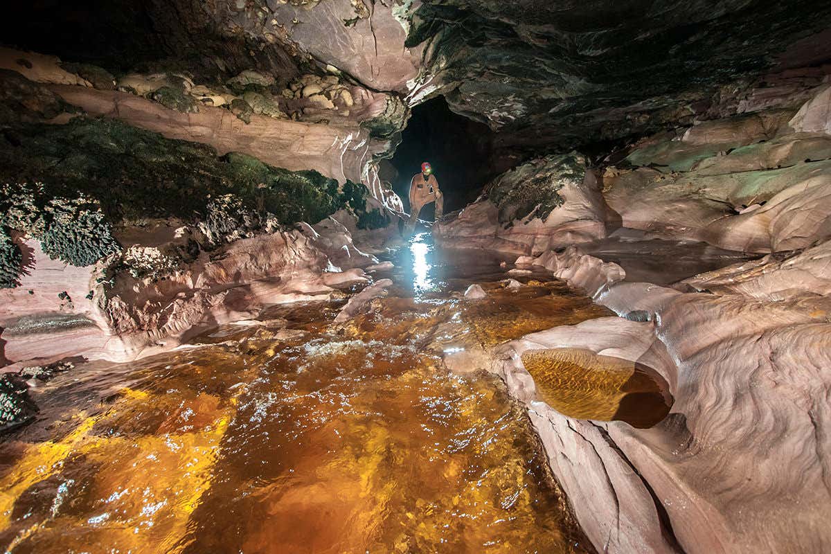 A cave chamber with pinkish orange rock and orange tinted water, with very irregular walls. It looks enchanting
