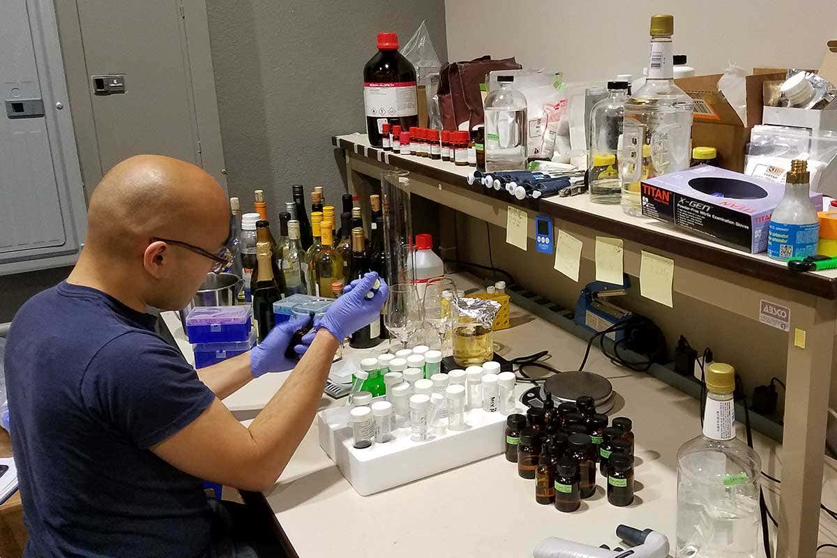 A man wearing gloves sits at a table covered in bottles containing different chemicals