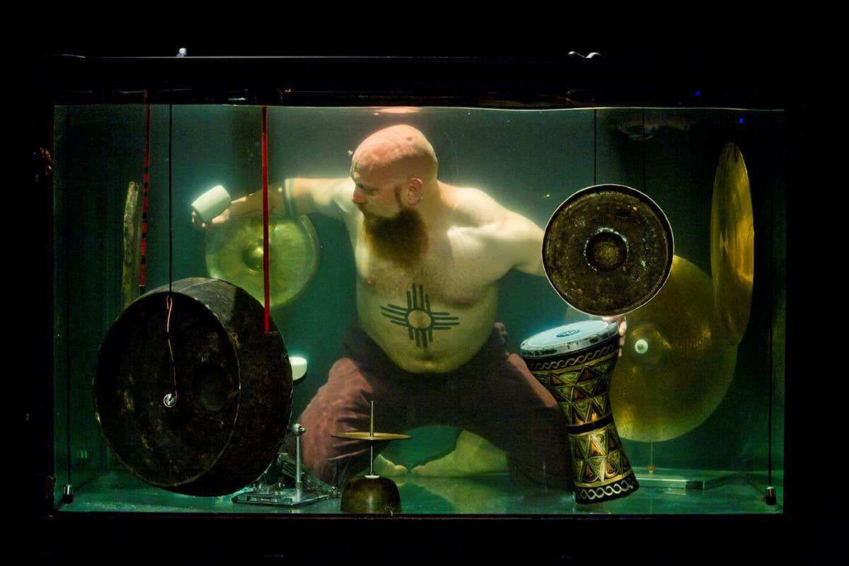 A big guy, barechested with a tattoo, bald head and beard, underwater, playing I think some cymbals