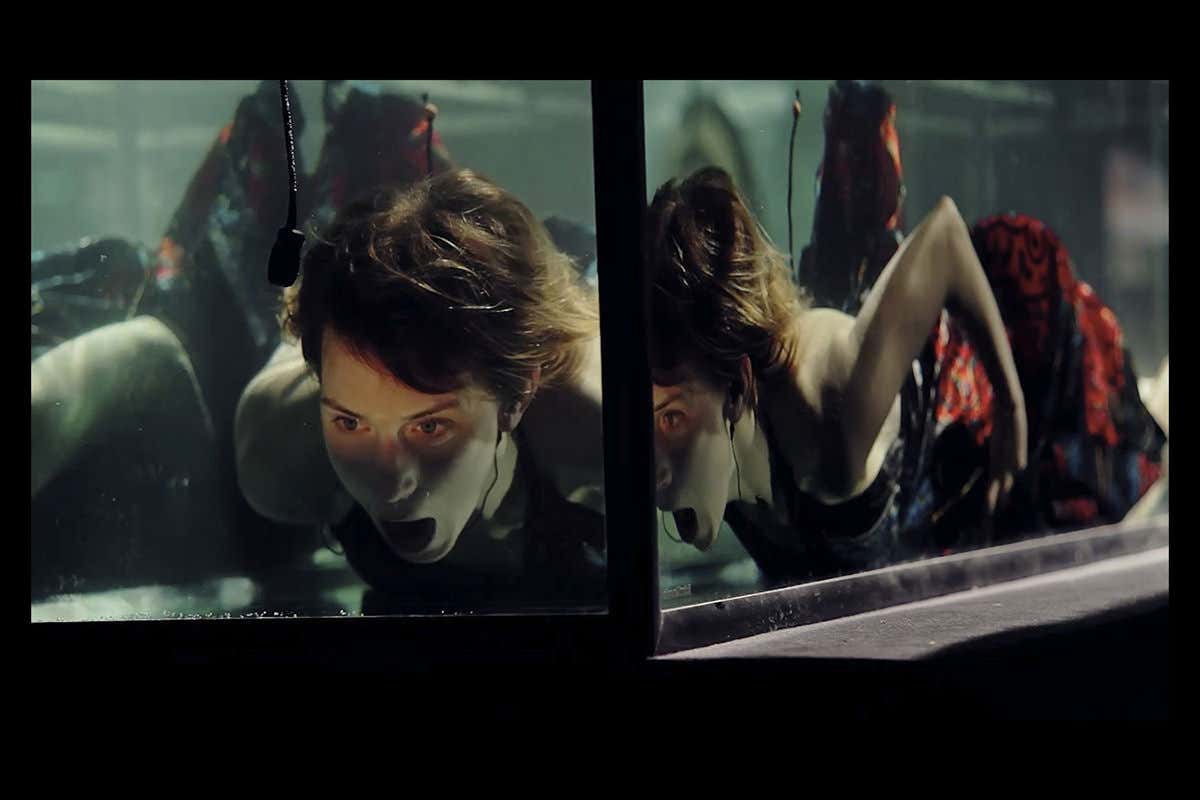 A woman - the singer in the band - lying on her front in a water tank, in a red dress, with her mouth open - presumably 