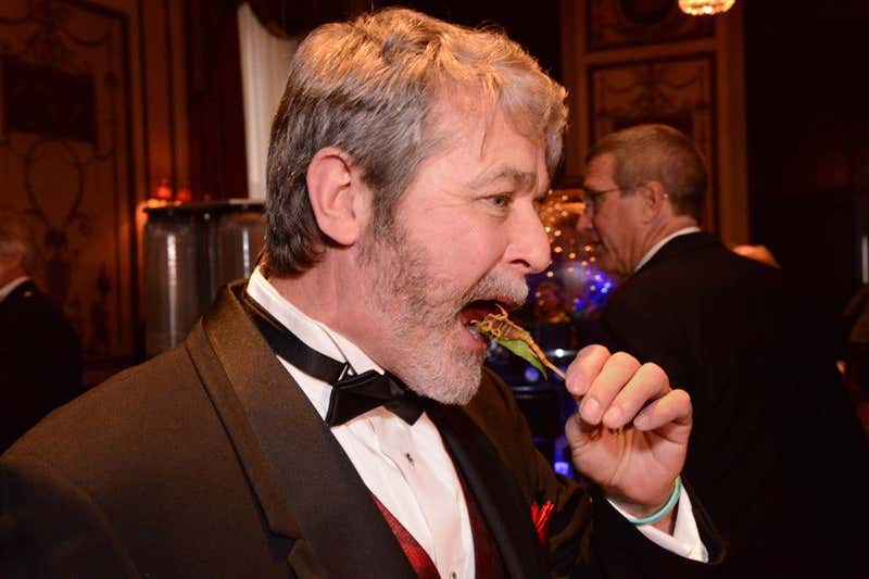 A man in black tie eating a scorpion on a stick