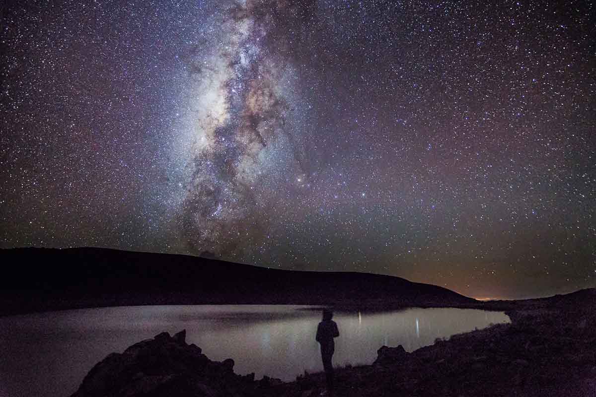 A landscape with a distant person looking up at the night sky where the Milky Way is visible