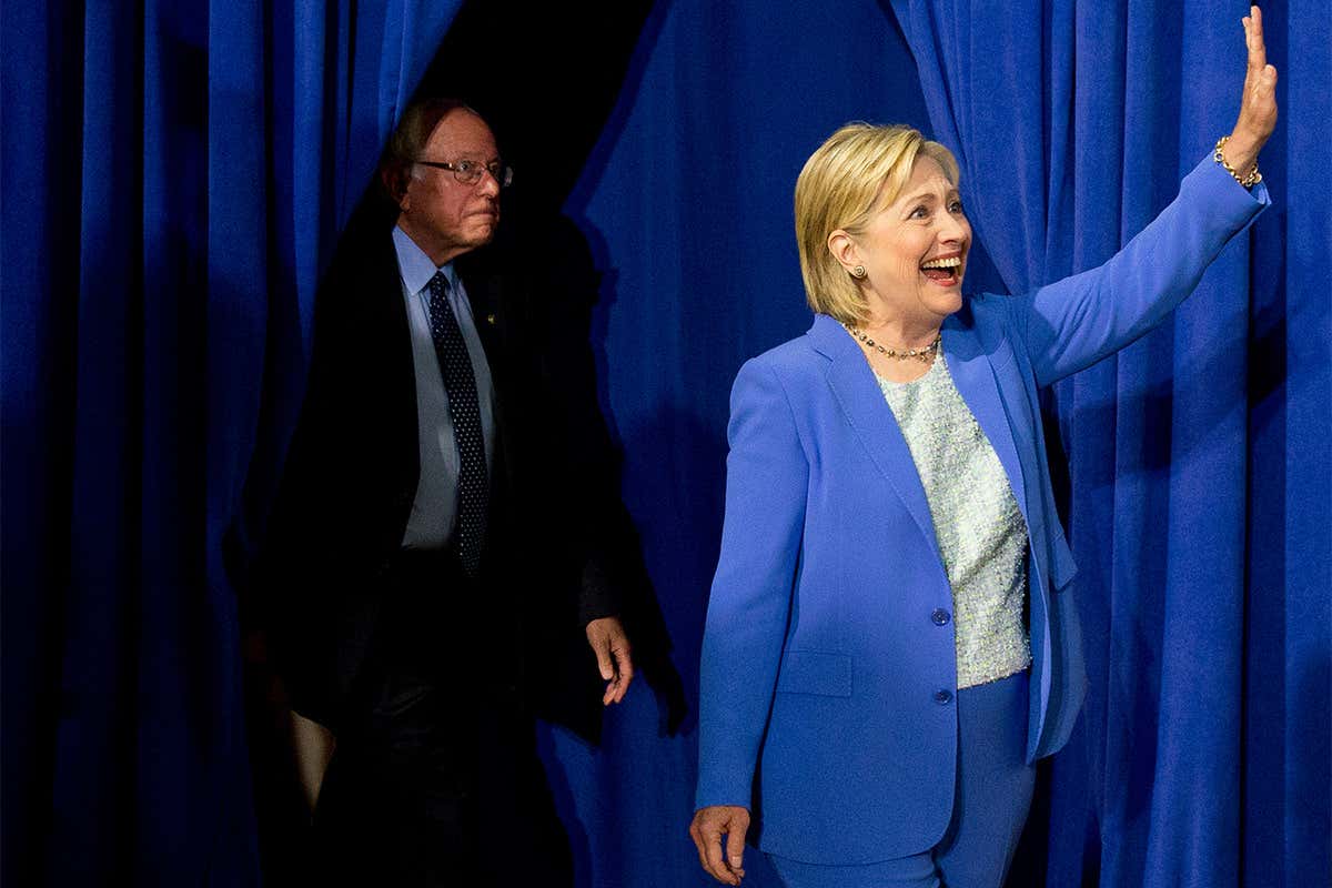Clinton waves in front of a stage curtain while Sanders emerges from the wings behind her