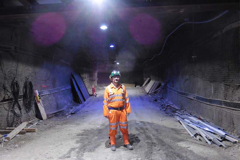 A man wearing an orange boiler suit, hard hat and headtorch stands in the mine