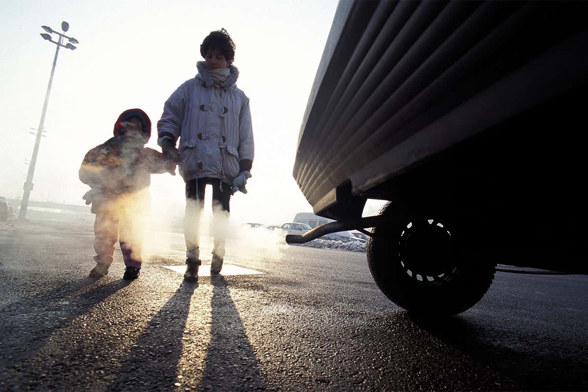 A car exhaust is pumping out gas while a person holding a child's hand can be seen in the background