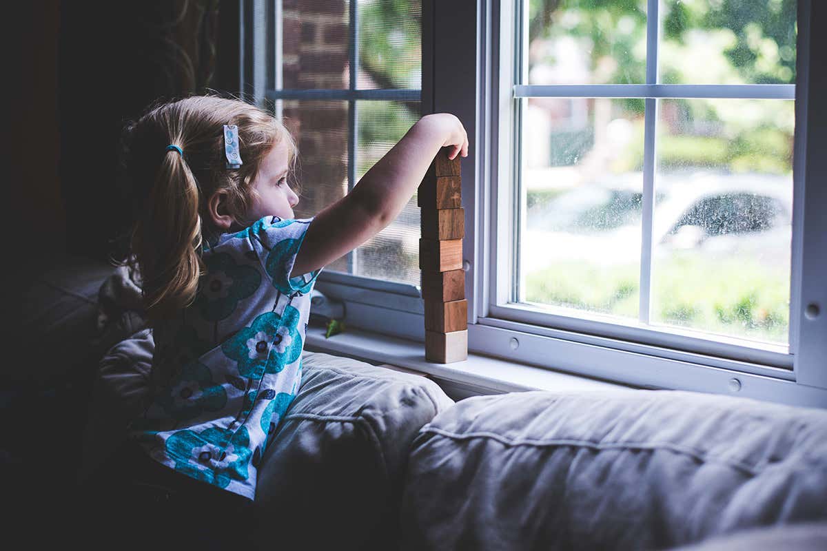 A little girl plays with wooden blocks, stacking them into a tower on a windowsill