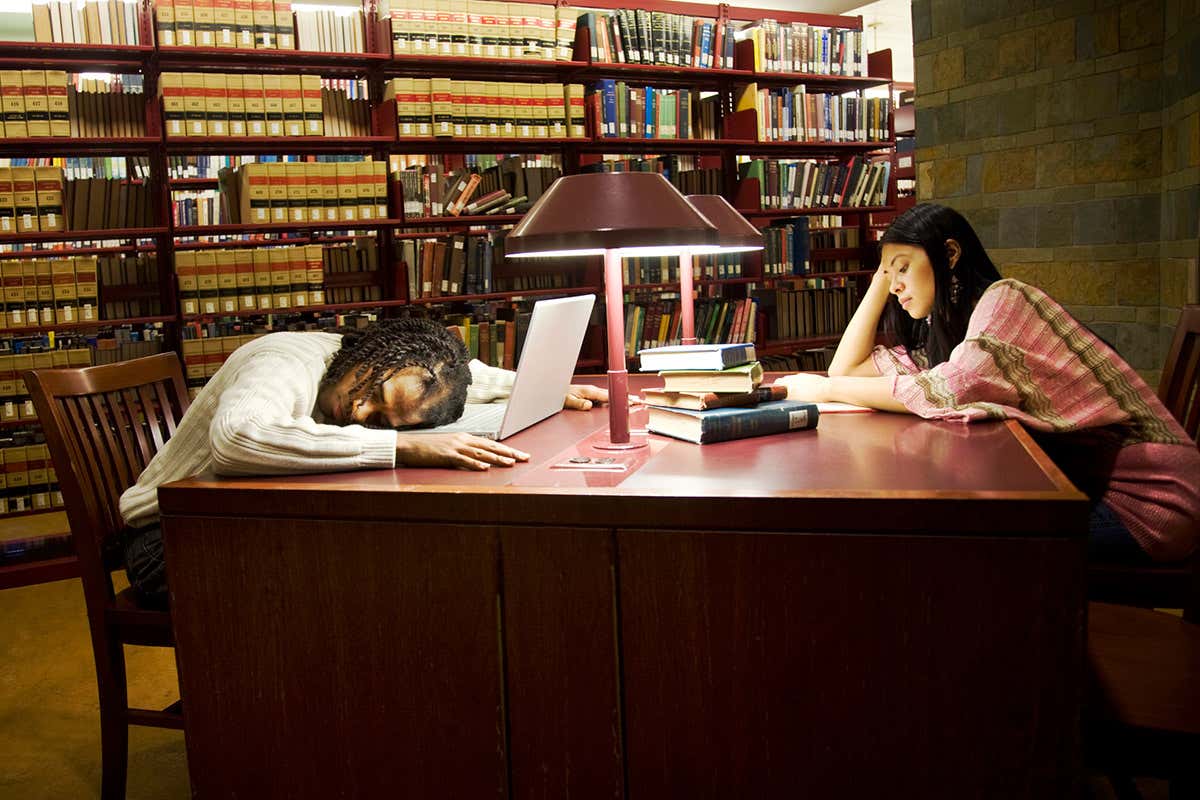 Two students in a library, one with head on the desk