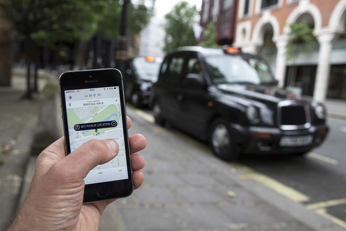 In the background, a regular black cab is parked beside the pavement of a suburban street, in the foreground a hand is holding a smartphone running the Uber app