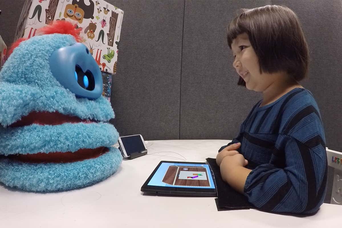 A child sits opposite a fluffy blue robot companion on a desk