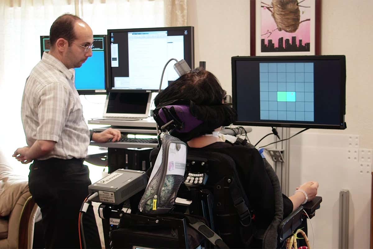 A person in a wheelchair looks towards a screen with a grid on it while a researcher looks on