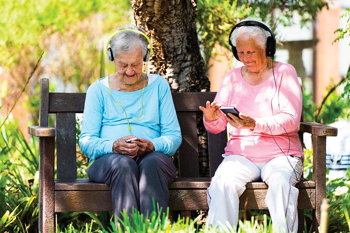 Two white-haired people on park bench with headphones on