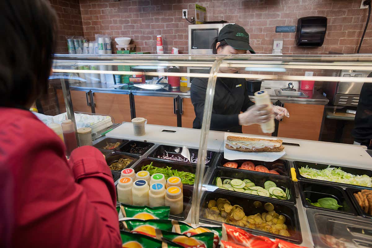 A person stands in front of the counter in a Subway restaurant as the server assembles a sandwich