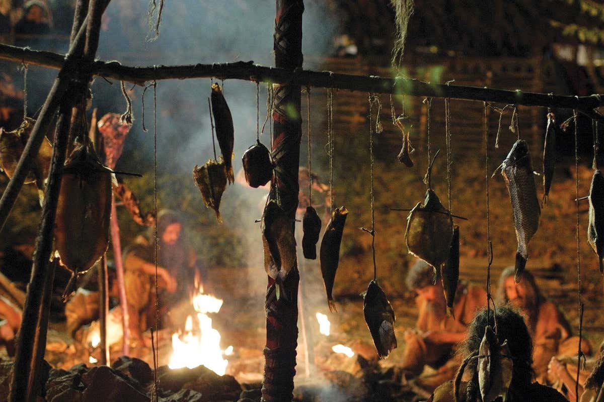 Neanderthals sat round a campfire with bits of meat hanging up in the foreground