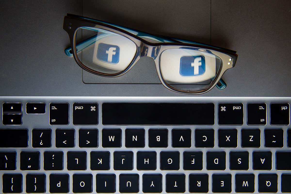 A pair of glasses next to a computer keyboard reflecting the Facebook logo