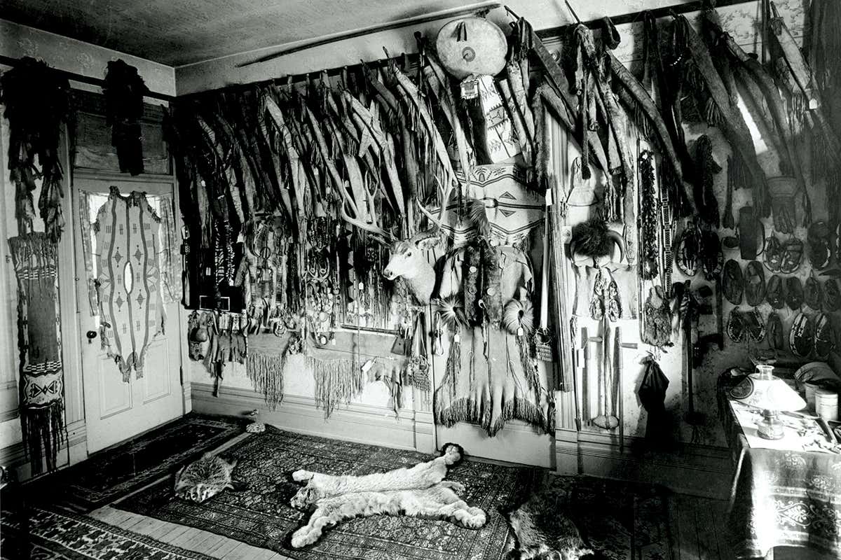 Black-and-white photo of room with Native America clothes and artefacts on the wall