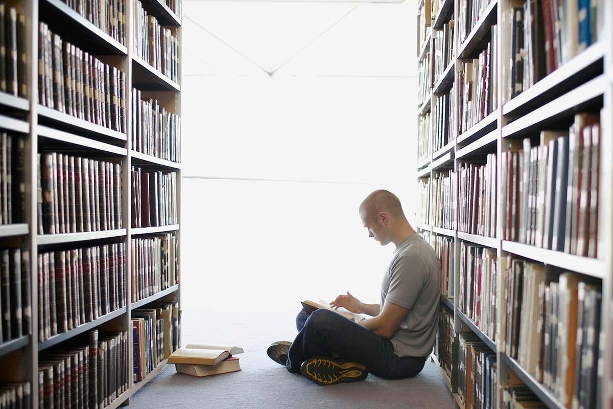 Man on floor reading between the stacks of a library shelf