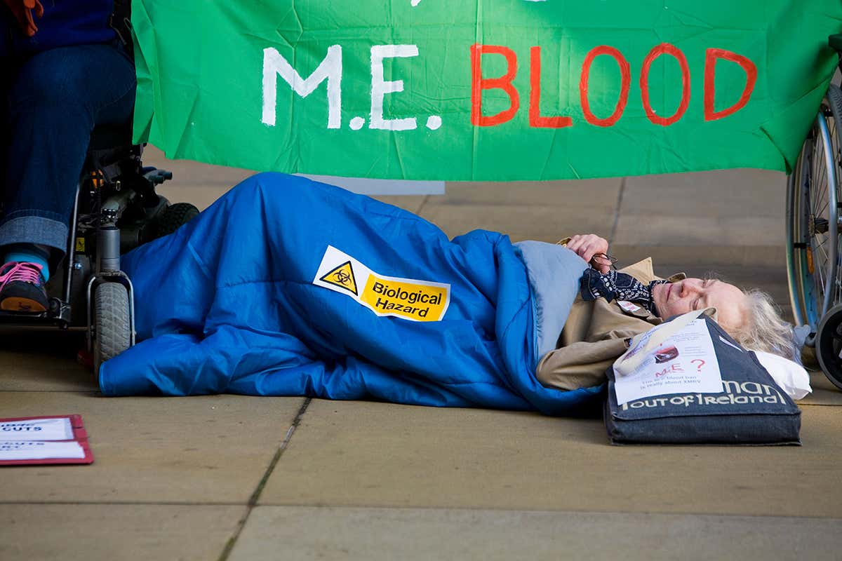 A person with ME lying on the floor with a biohazard sticker on her. A green banner behind with ME Blood written on it.