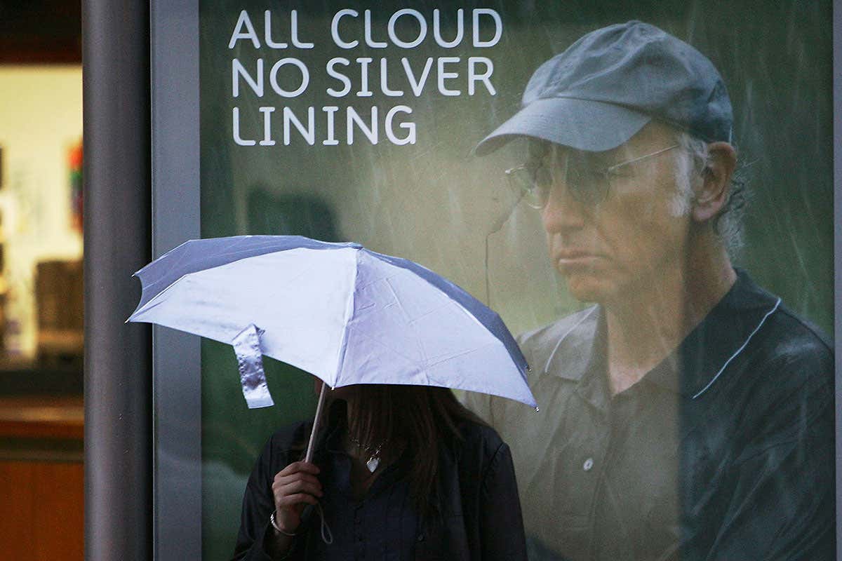 A person with an umbrella standing in the rain in front of an advert that says 