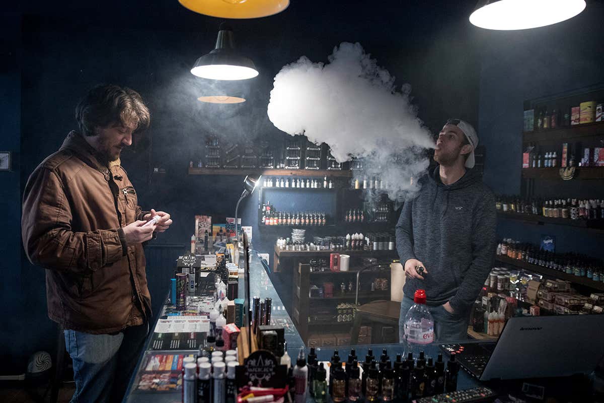 Two men standing in a vape shop, one inspecting products, the other behind the counter exhaling smoke