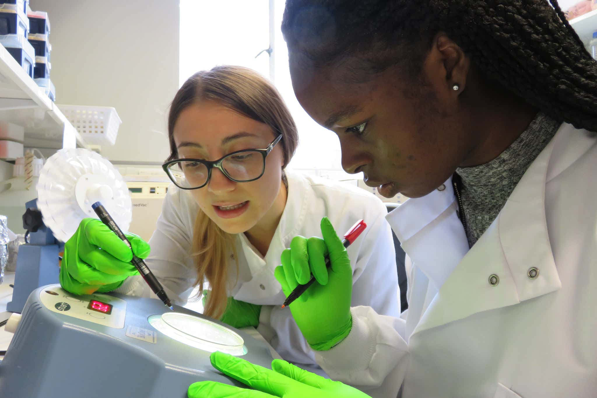 Two young women in a lab wearing white coats and holding pens