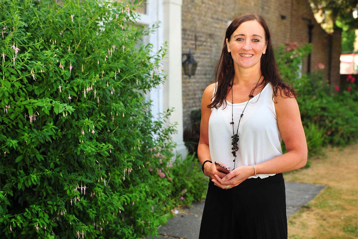 Portrait of Kath Sansom, a young, lively-looking woman, outside a house