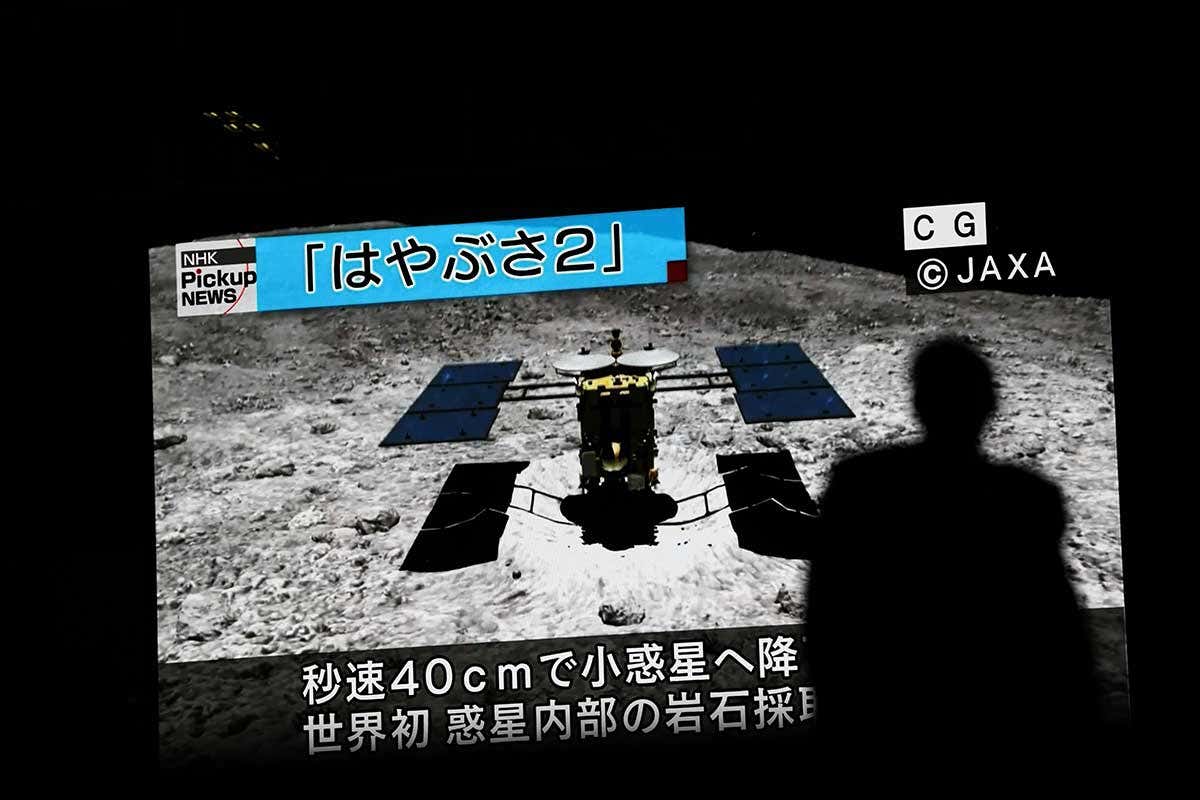In this picture taken on July 10, 2019 a man walks past a screen displaying a computer-generated image of the Hayabusa2 probe, during a news broadcast at Akihabara district in Tokyo. - Japan's Hayabusa2 probe landed successfully on a distant asteroid for a final touchdown on July 11, 2019, hoping to collect samples that could shed light on the evolution of the solar system