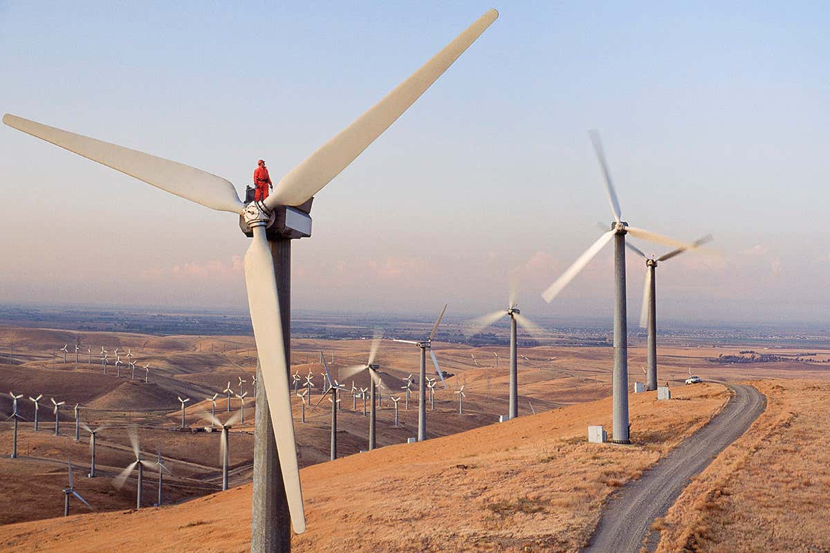High angle view of a male worker standing atop a wind turbine at dusk wearing protective work clothes and safety hardhat inspecting machinery