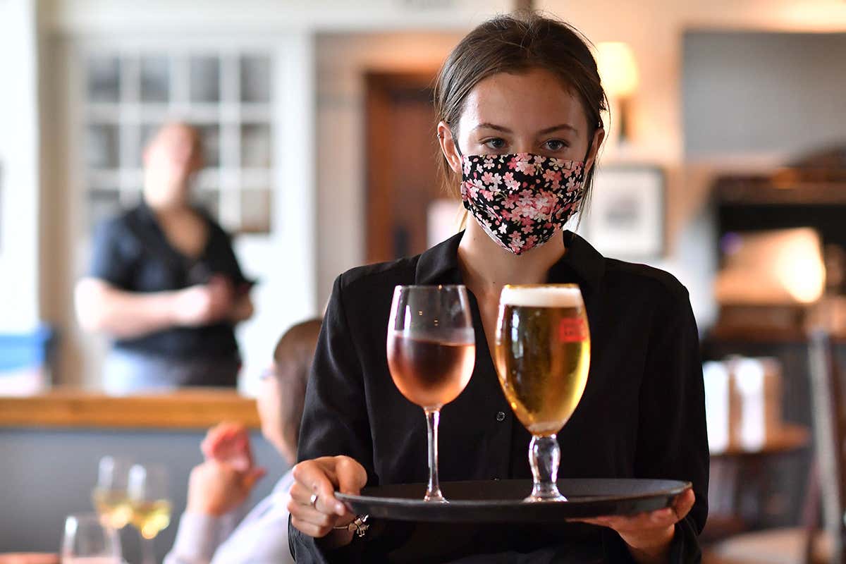 A restaurant staff member wearing a mask carries a tray with drinks
