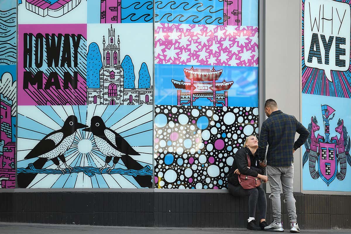 Two people face each other, one sitting and one standing, near artwork in Newcastle city centre, north-east England