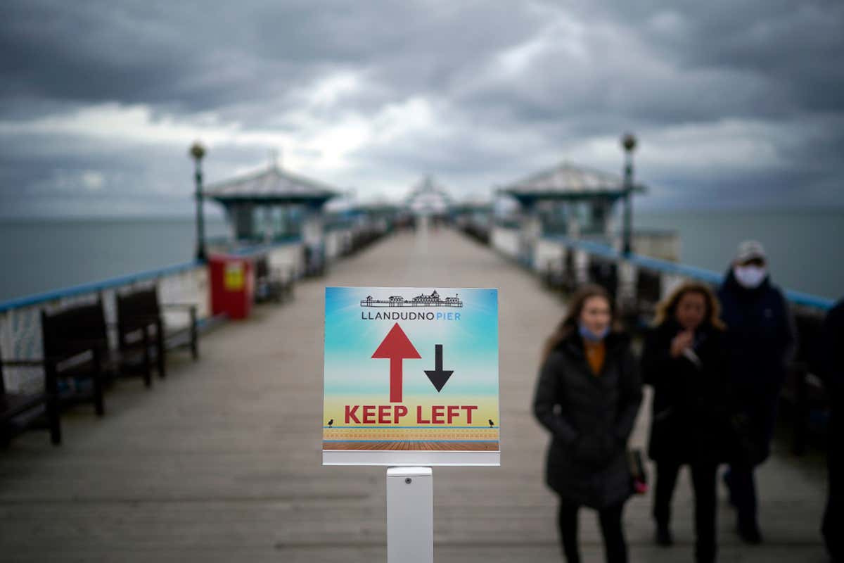 People walk on Llandudno Pier, Conwy, Wales beside a sign that reads: 'Llandudno Pier, Keep left'