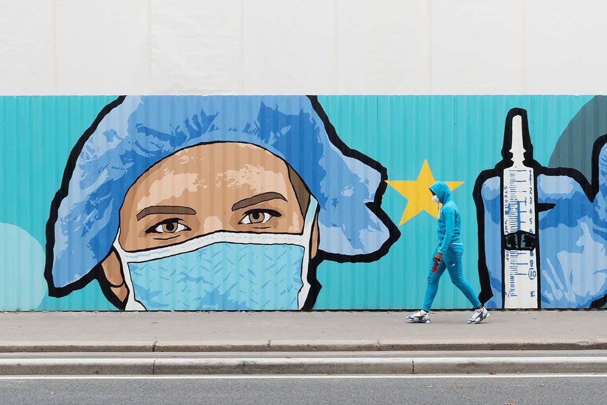 Graffiti showing a person wearing personal protective equipment and holding a syringe in their gloved hand on the wall, with a person walking in front of it.