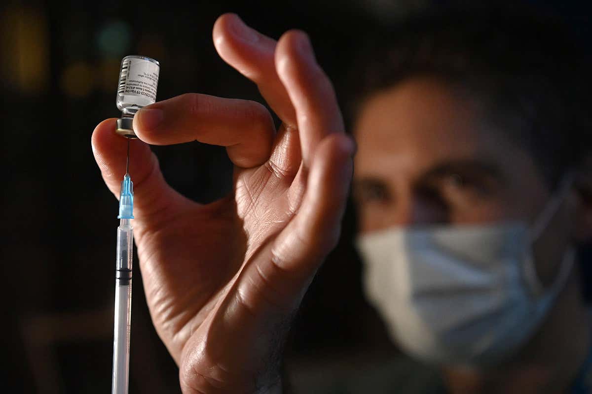 A person wearing personal protective equipment holds a vaccine vial and syringe between their fingers
