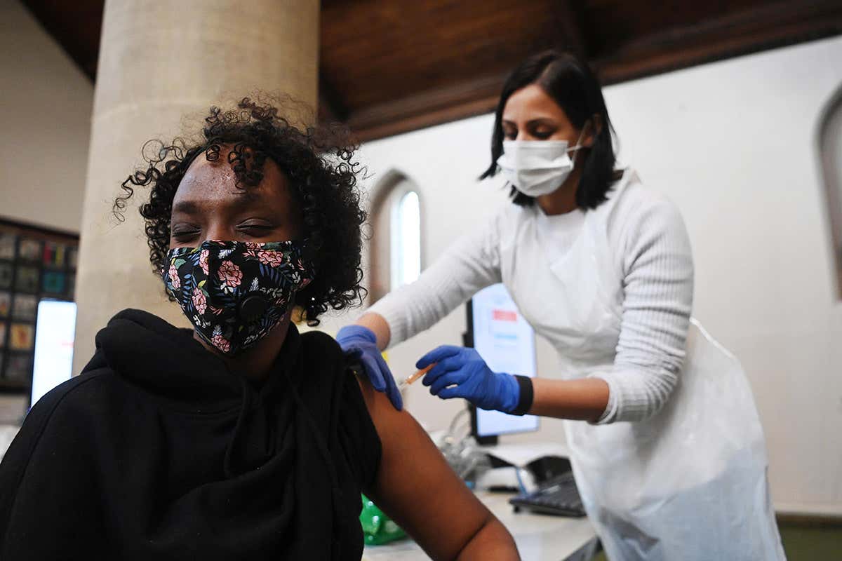 A healthcare worker wearing personal protective equipment administers an injection to a person wearing a face covering