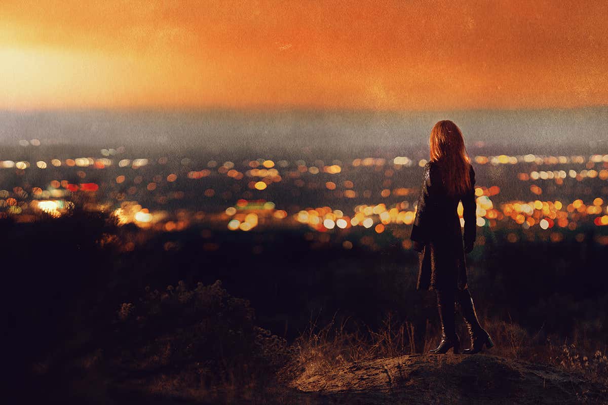 Back view of woman with long red hair wearing black coat and heeled boots standing on hillside looking at sunset over valley of city lights