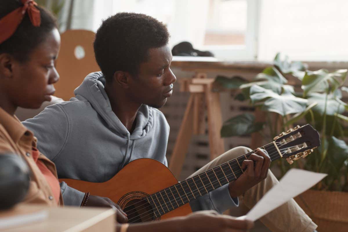 Side view portrait of two young African-American musicians playing guitar and writing music together while sitting on floor in recording studio, copy space