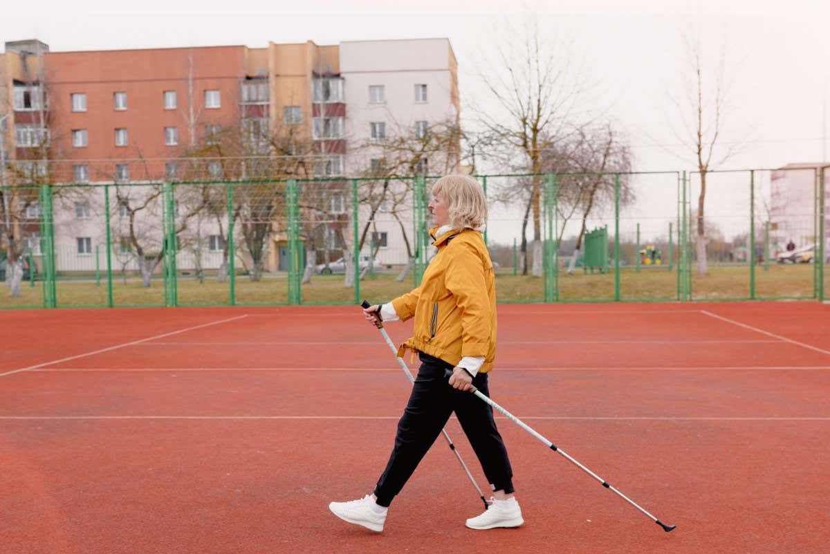 2F7DHH7 An elderly woman in a yellow sports jacket practices Nordic walking outdoors on the stadium's rubber treadmill. A sunny sunset. Older women walk with