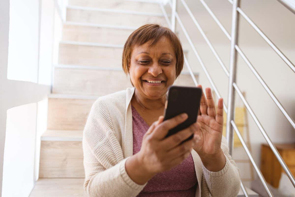 Happy senior woman waving hand at video call through smart phone while sitting on steps at home