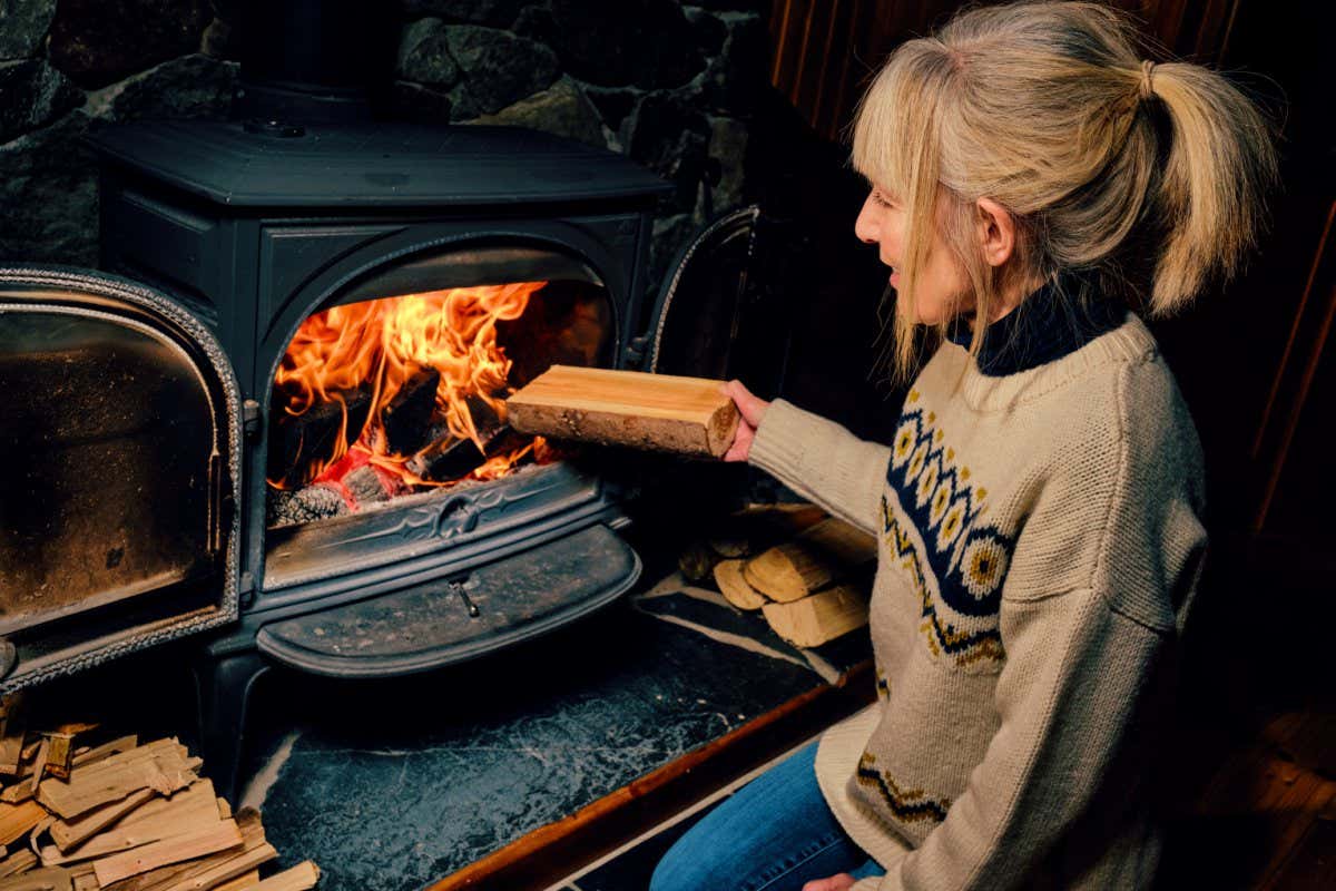 Woman putting a well dried log into a glowing wood burning stove.