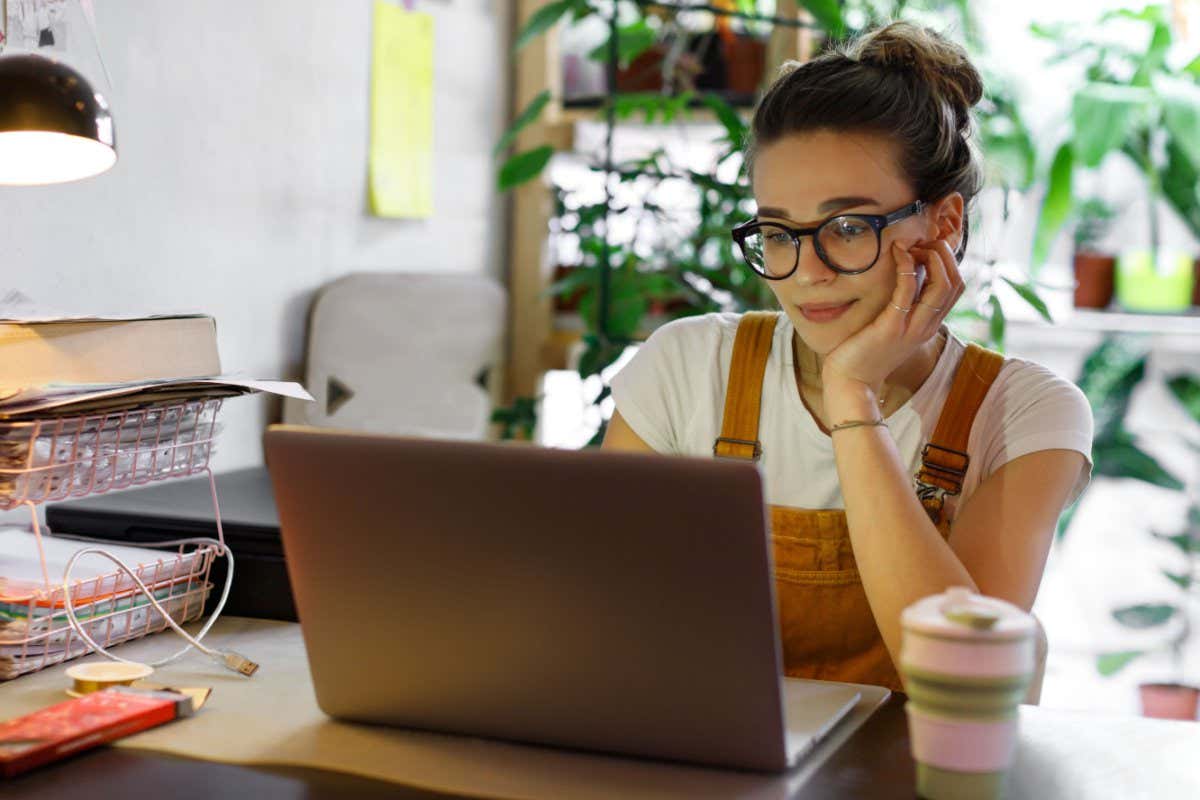 Young female gardener in glasses using laptop, communicates on internet with customer in home garden/greenhouse, reusable coffee/tea mug on table.Cozy office workplace, remote work, E learning concept; Shutterstock ID 1674467302; purchase_order: -; job: -; client: -; other: -